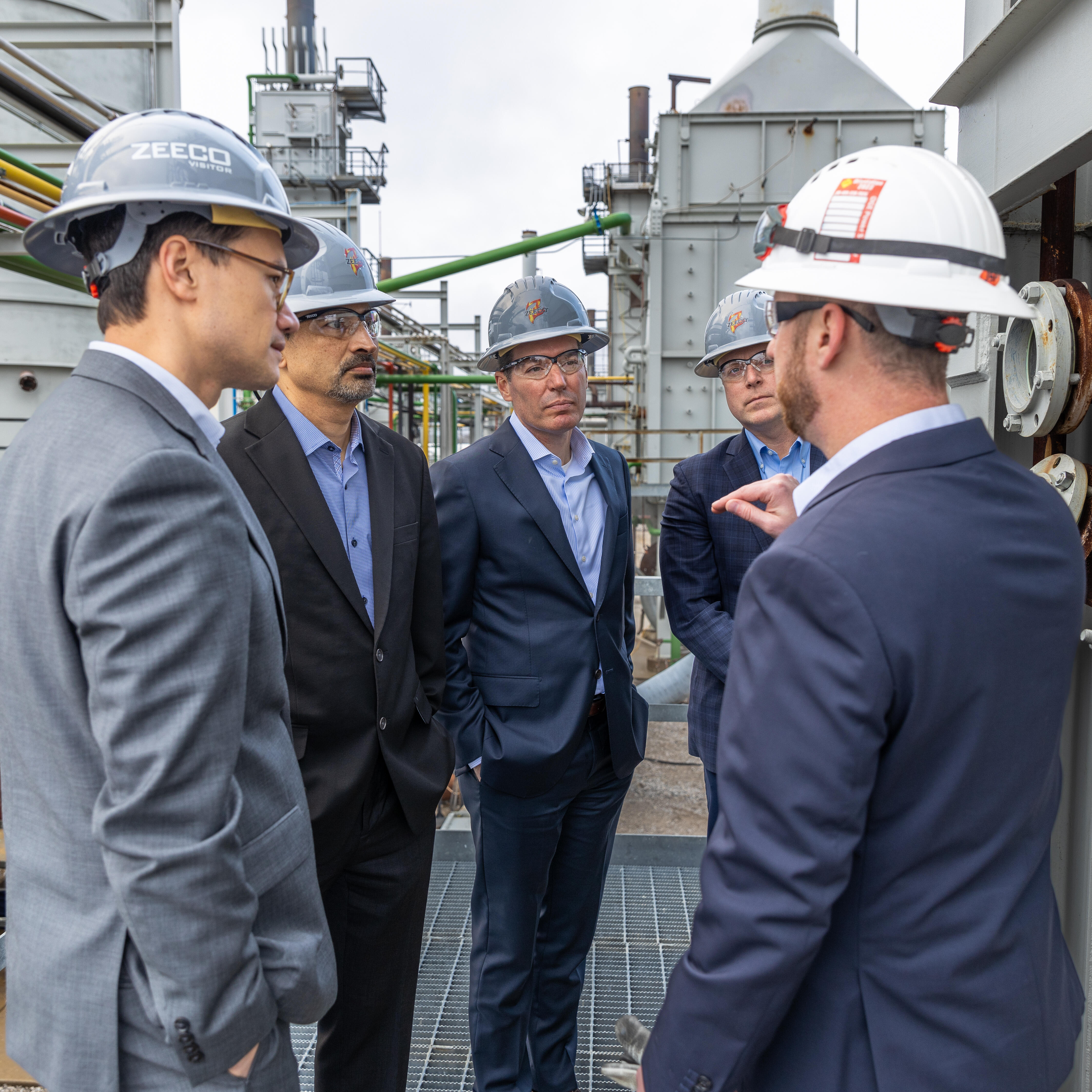 Five men talking in a group, wearing suits and Zeeco helmets.