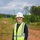Worker in high-visibility vest and hard hat walking through a wooded area on a dirt trail.