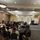 A conference or seminar is taking place in a large banquet-style room with round tables and black tablecloths. Attendees are seated and facing a stage where a speaker stands at a podium beside floral arrangements and U.S. and state flags.