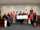 A group of about fourteen people pose indoors in a meeting room, standing in a line against a light-colored wall. Several of them hold an oversized check made out to “Magnolia School District” for $20,000.