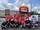 A large group of people pose in front of a parade float decorated with a colorful cluster of balloons and a hand‑painted banner that reads “ExxonMobil.” Most of the group wear matching red “ExxonMobil Community Partners” T‑shirts, while others in front wear black shirts and cowboy hats.