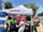 A small outdoor booth is set up under a white tent with the ExxonMobil logo. Several people are gathered around a table with brochures, giveaway items, and a display case, while three staff members in red ExxonMobil shirts talk with visitors. A blue informational sign about “Mobil Lithium” stands on the left