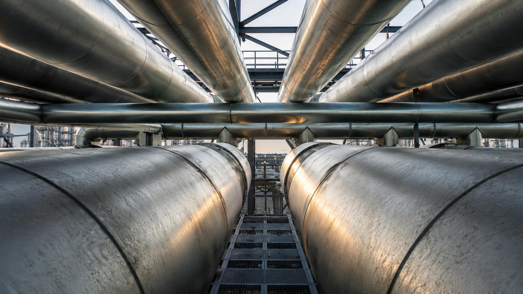 A low-angle view shows a network of large industrial pipes running parallel above and beside a grated walkway. The metallic surfaces catch soft light, creating warm reflections along the cylindrical forms.