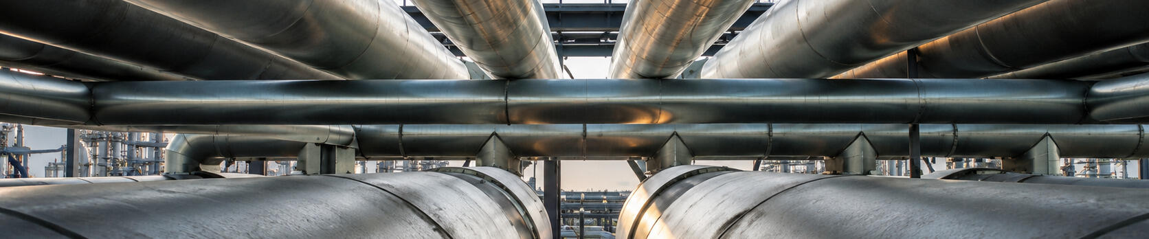 A low-angle view shows a network of large industrial pipes running parallel above and beside a grated walkway. The metallic surfaces catch soft light, creating warm reflections along the cylindrical forms.