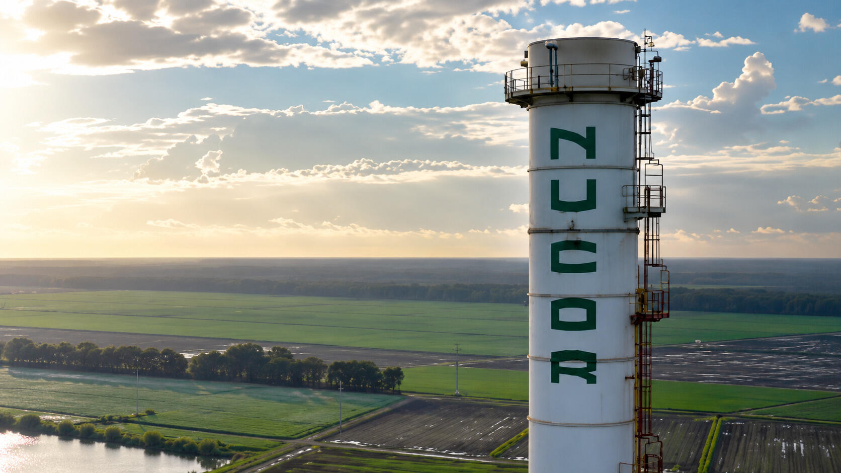 A tall industrial smokestack stands in the foreground with the word “NUCOR” painted vertically in large green letters.